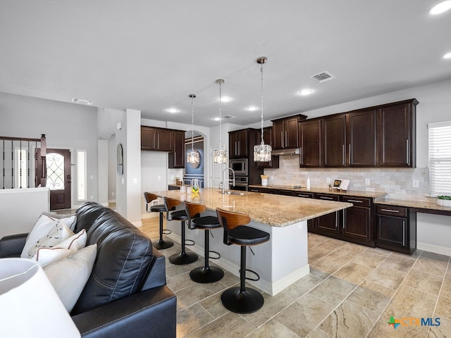 kitchen featuring light stone countertops, a kitchen bar, a kitchen island with sink, pendant lighting, and decorative backsplash