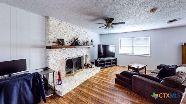 living room featuring a textured ceiling, hardwood / wood-style flooring, a stone fireplace, and ceiling fan