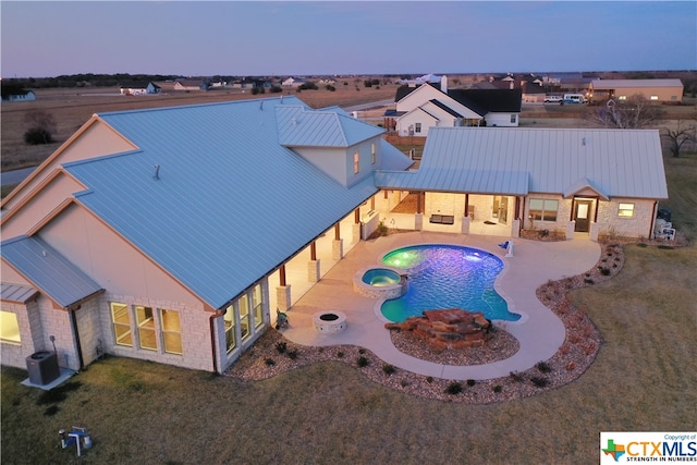 pool at dusk featuring a patio area, central AC, a lawn, and an in ground hot tub