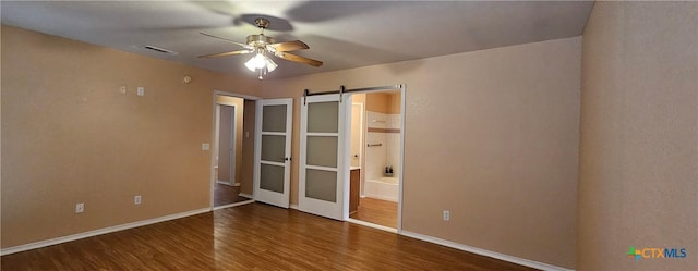 unfurnished bedroom with ensuite bathroom, a barn door, dark hardwood / wood-style floors, and ceiling fan