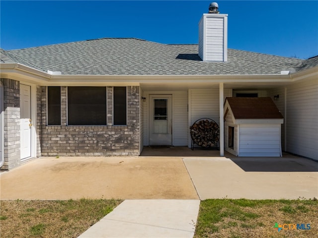 property entrance with a patio area, brick siding, roof with shingles, and a chimney