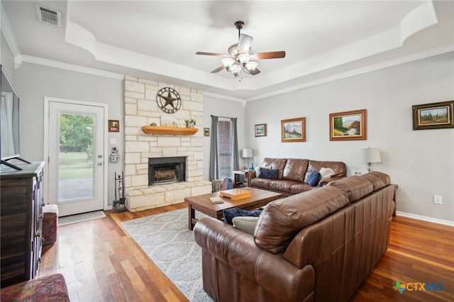 living room featuring wood-type flooring, a raised ceiling, a stone fireplace, and crown molding