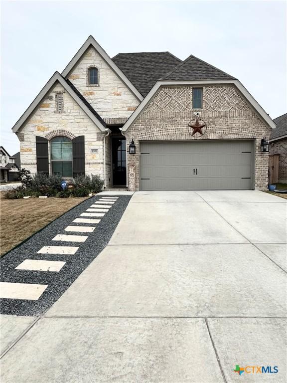 french country inspired facade featuring an attached garage, stone siding, concrete driveway, and brick siding