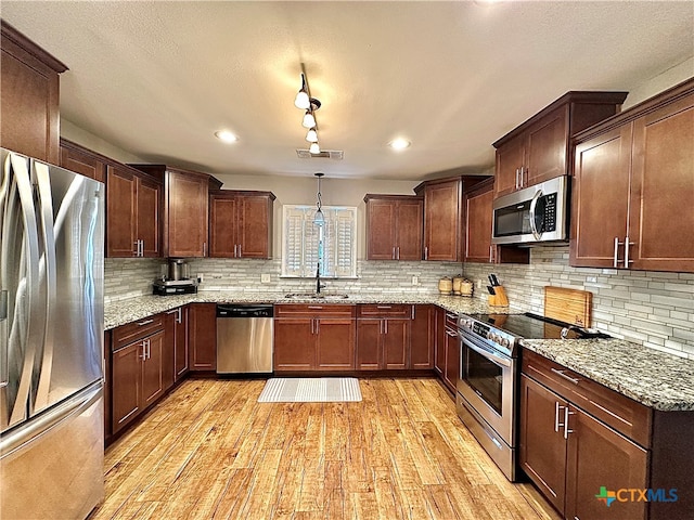 kitchen with light wood-type flooring, appliances with stainless steel finishes, pendant lighting, light stone countertops, and sink