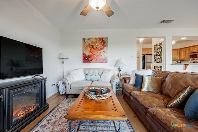 living area featuring visible vents, a glass covered fireplace, light wood-style floors, baseboards, and ceiling fan