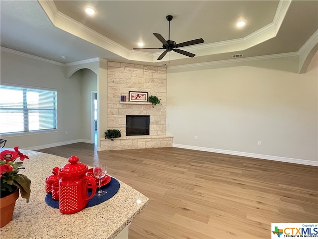 living room with a stone fireplace, hardwood / wood-style floors, ornamental molding, ceiling fan, and a raised ceiling