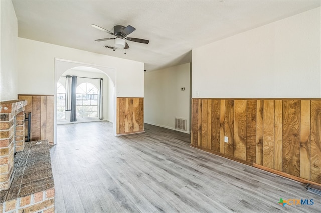 unfurnished living room featuring a brick fireplace, ceiling fan, wooden walls, and light hardwood / wood-style flooring