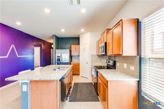 kitchen with sink, appliances with stainless steel finishes, light stone countertops, backsplash, and an island with sink