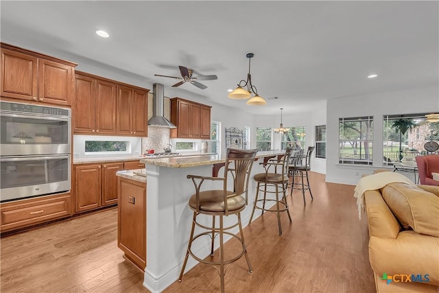 kitchen featuring recessed lighting, stainless steel double oven, a breakfast bar, wall chimney range hood, and open floor plan