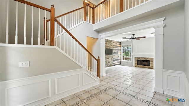 staircase featuring a stone fireplace, a ceiling fan, a decorative wall, wainscoting, and tile patterned flooring