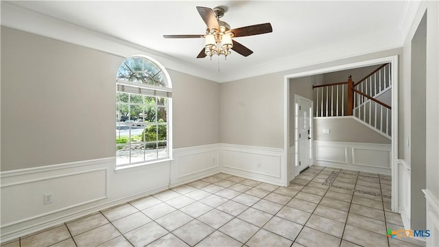 empty room with ceiling fan, stairs, a wainscoted wall, crown molding, and a decorative wall
