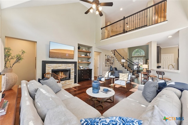 living area with a ceiling fan, wood finished floors, stairway, a towering ceiling, and a stone fireplace