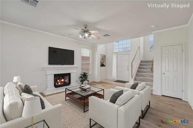 living area featuring visible vents, light wood finished floors, a lit fireplace, stairs, and crown molding