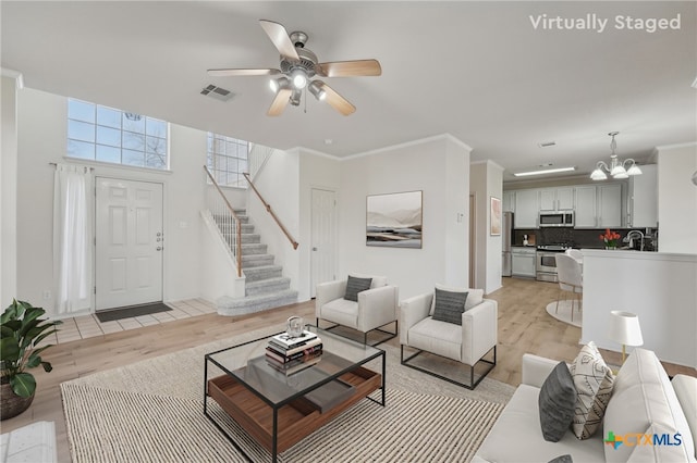 living room featuring light wood finished floors, visible vents, crown molding, stairway, and ceiling fan with notable chandelier