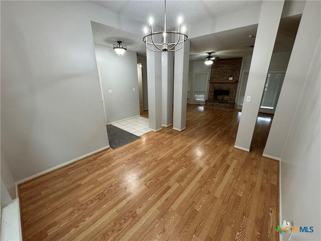 unfurnished dining area with a textured ceiling, ceiling fan with notable chandelier, a fireplace, baseboards, and light wood-style floors