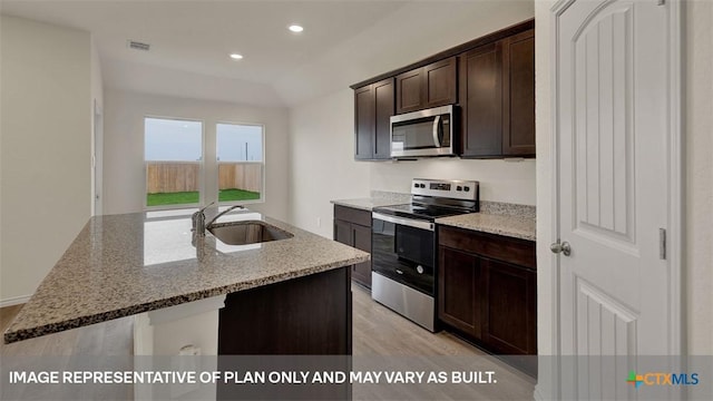 kitchen with sink, appliances with stainless steel finishes, dark brown cabinets, light stone countertops, and an island with sink