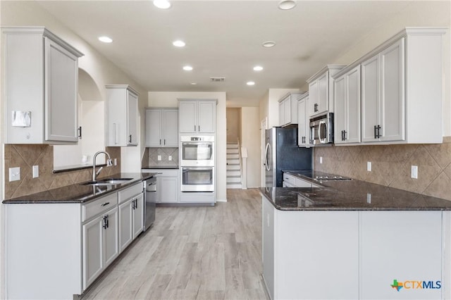 kitchen with stainless steel appliances, a peninsula, a sink, light wood finished floors, and dark stone countertops