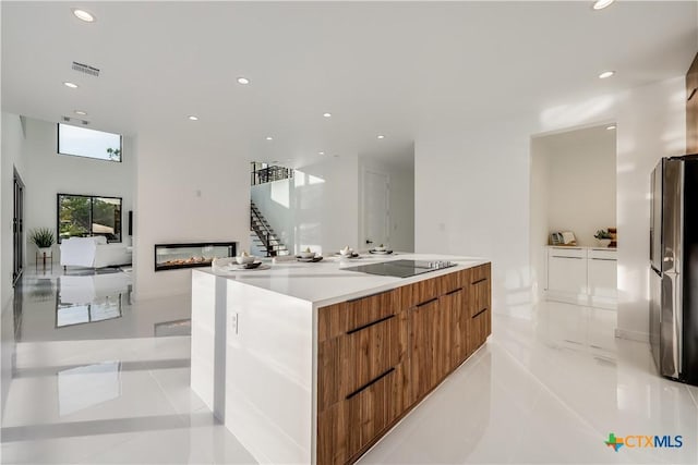 kitchen with stainless steel refrigerator with ice dispenser, black electric stovetop, a spacious island, light tile patterned floors, and a multi sided fireplace