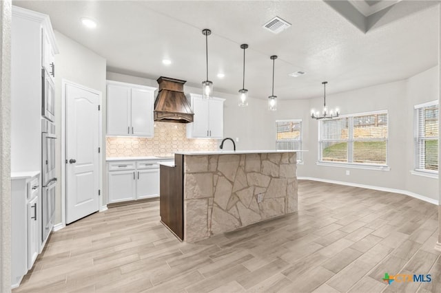 kitchen with pendant lighting, tasteful backsplash, white cabinetry, custom range hood, and a center island with sink