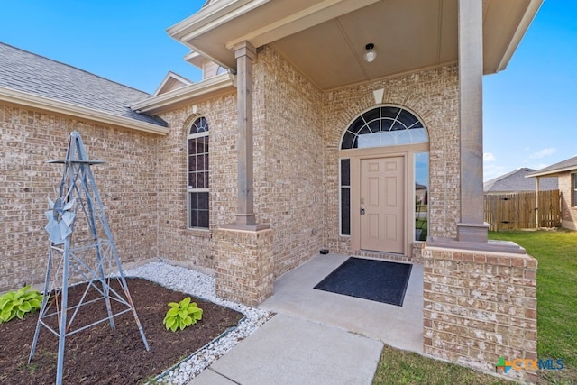 view of exterior entry featuring brick siding, roof with shingles, and fence