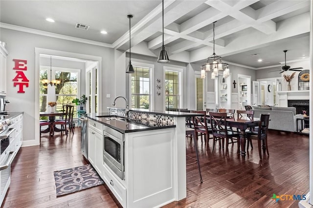 kitchen featuring white cabinetry, a center island with sink, stainless steel microwave, decorative light fixtures, and ceiling fan with notable chandelier