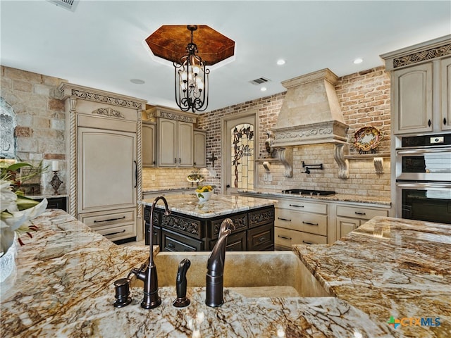 kitchen with backsplash, light stone counters, custom range hood, stainless steel double oven, and cream cabinetry