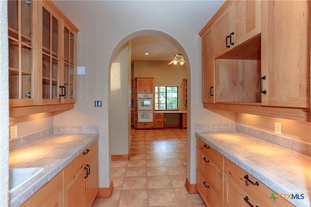 kitchen with tile counters, sink, ceiling fan, double oven, and light brown cabinets