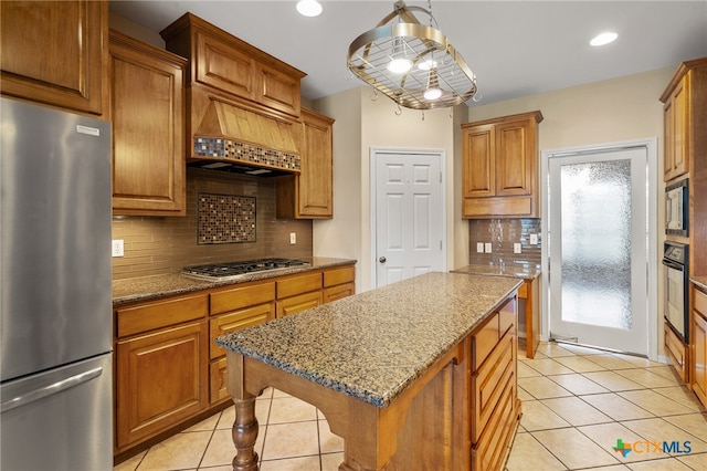 kitchen featuring custom exhaust hood, a kitchen island, light stone countertops, appliances with stainless steel finishes, and decorative light fixtures