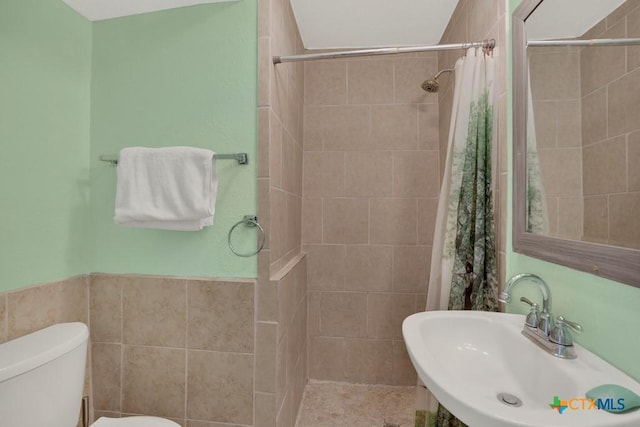 full bath with tiled shower, wainscoting, toilet, a sink, and tile walls