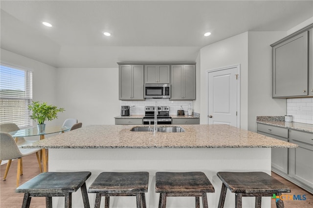 kitchen featuring sink, a center island with sink, gray cabinets, stainless steel appliances, and light stone countertops