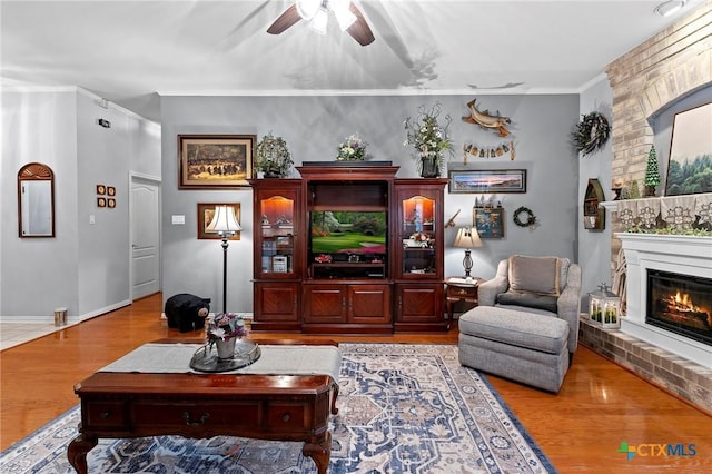 living room with a brick fireplace, ornamental molding, ceiling fan, and light wood-type flooring