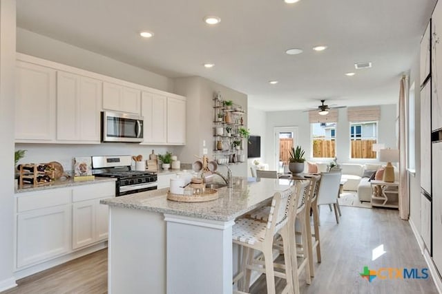 kitchen with white cabinetry, light wood-type flooring, appliances with stainless steel finishes, light stone countertops, and a kitchen island with sink