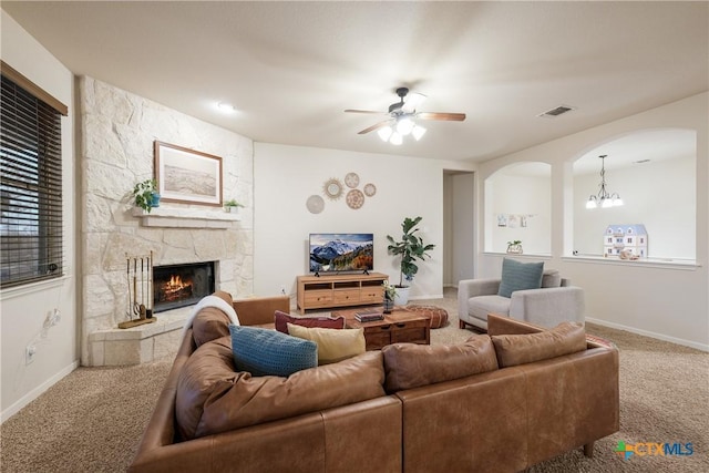 carpeted living room featuring a stone fireplace and ceiling fan with notable chandelier