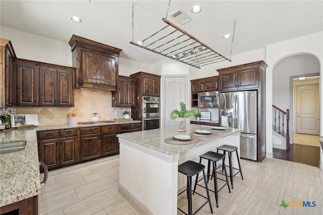 kitchen featuring light stone countertops, stainless steel appliances, tasteful backsplash, dark brown cabinets, and a kitchen island