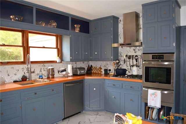 kitchen with stainless steel appliances, wooden counters, wall chimney range hood, sink, and blue cabinets