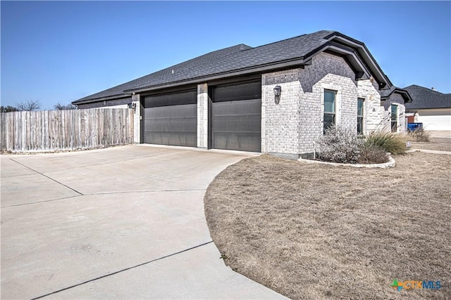 view of side of home featuring brick siding, a shingled roof, an attached garage, fence, and driveway