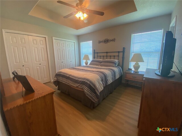 bedroom with a ceiling fan, light wood finished floors, multiple closets, and a tray ceiling