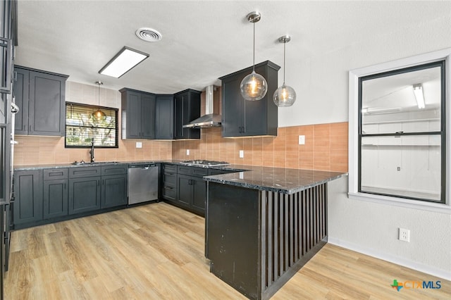 kitchen featuring wall chimney range hood, stainless steel appliances, dark stone countertops, hanging light fixtures, and light hardwood / wood-style flooring