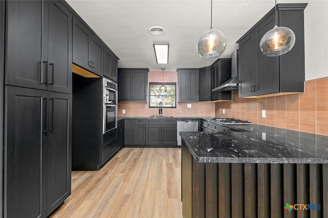 kitchen featuring appliances with stainless steel finishes, light wood-type flooring, dark stone counters, pendant lighting, and sink