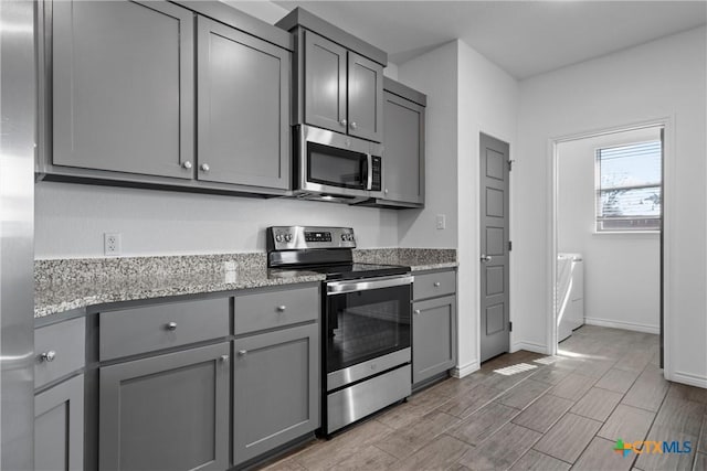 kitchen with gray cabinetry, light stone counters, appliances with stainless steel finishes, baseboards, and washing machine and clothes dryer