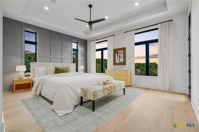 bedroom featuring multiple windows, light hardwood / wood-style floors, a raised ceiling, and ceiling fan