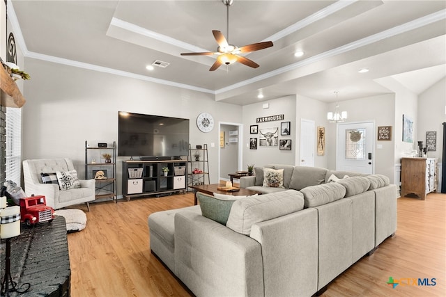 living room with a tray ceiling, visible vents, light wood-style flooring, ornamental molding, and ceiling fan with notable chandelier