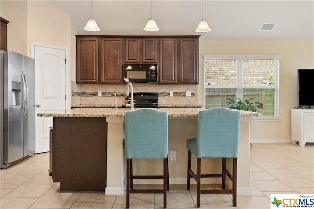 kitchen with light stone countertops, backsplash, stainless steel appliances, hanging light fixtures, and an island with sink