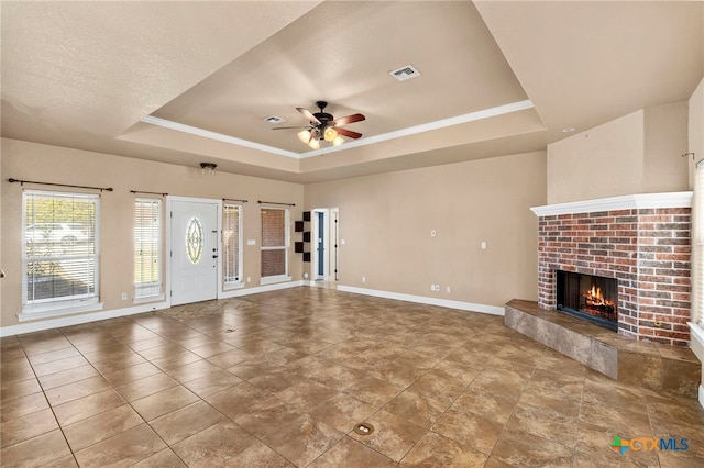 unfurnished living room featuring a brick fireplace, a textured ceiling, ornamental molding, a tray ceiling, and ceiling fan