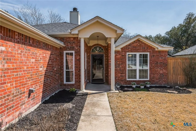 entrance to property featuring a shingled roof, a lawn, a chimney, fence, and brick siding