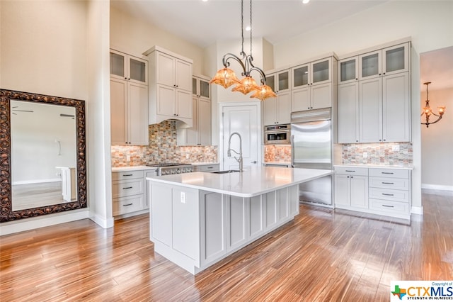 kitchen with sink, a kitchen island with sink, light wood-type flooring, appliances with stainless steel finishes, and decorative light fixtures