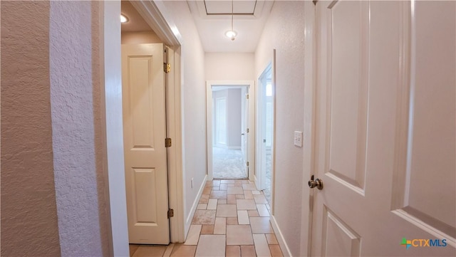 hallway featuring a textured wall, stone finish flooring, baseboards, and attic access