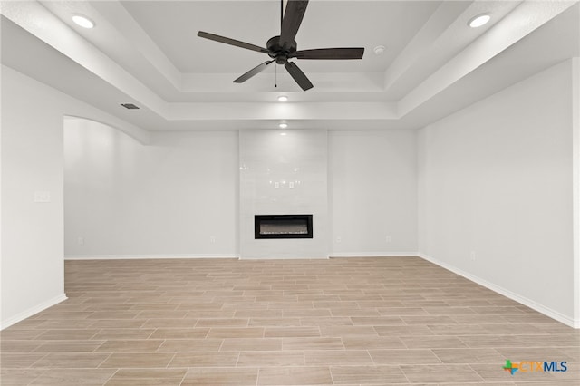 unfurnished living room featuring light wood-type flooring, a large fireplace, ceiling fan, and a tray ceiling