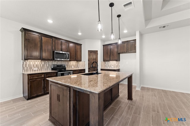 kitchen featuring a center island with sink, light stone counters, sink, pendant lighting, and appliances with stainless steel finishes