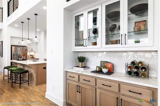 bar with stainless steel refrigerator with ice dispenser, backsplash, light stone counters, light hardwood / wood-style flooring, and hanging light fixtures